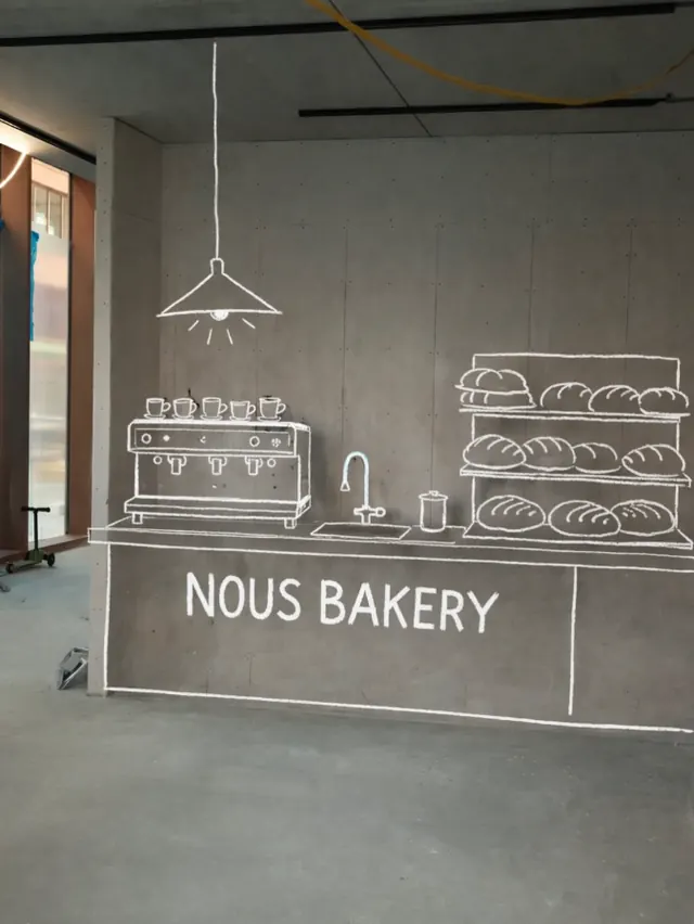 A chalk drawing of a bakery counter, espresso machine and shelves of bread on a raw concrete wall, captioned NOUS BAKERY.