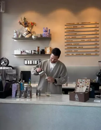 A barista at nous pouring steamed milk, with the wooden letterboard menu on the wall behind.