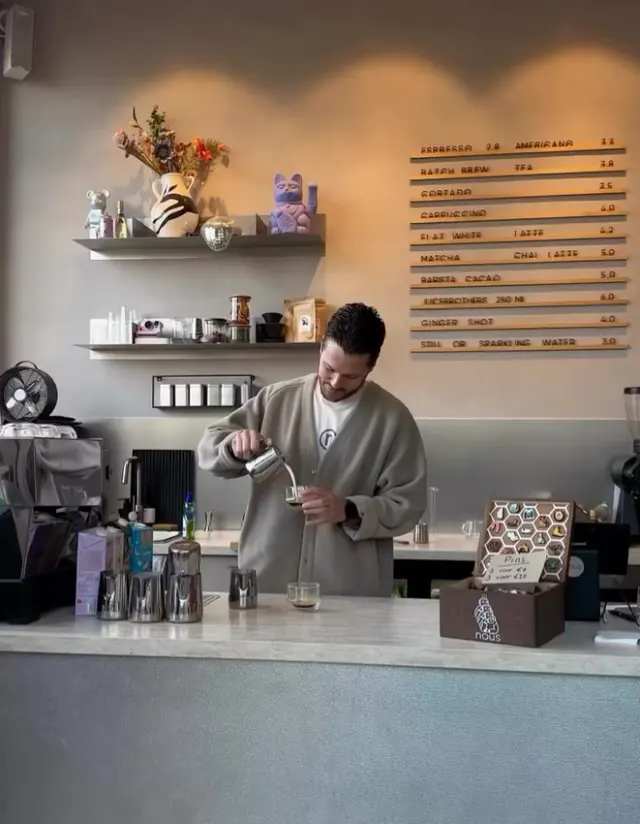 A barista pouring milk behind the pale counter at nous, under the wooden letterboard menu.