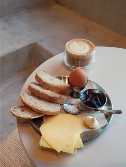 A nous breakfast plate: sliced bread, a boiled egg, cheese, butter and jam beside a cappuccino.