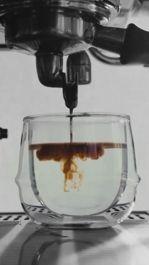 Espresso extracting from a bare portafilter into a clear glass cup, the shot blooming through hot water.