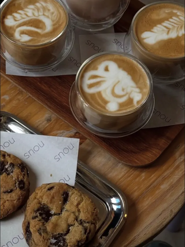 Three glass cups of latte art on a wooden tray beside sea salt chocolate chip cookies on nous napkins.