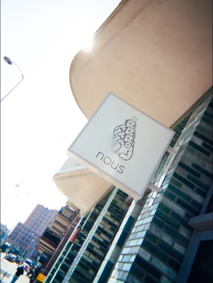 The hanging nous sign in the sun, showing three stacked figures each holding a cup above the lowercase wordmark.