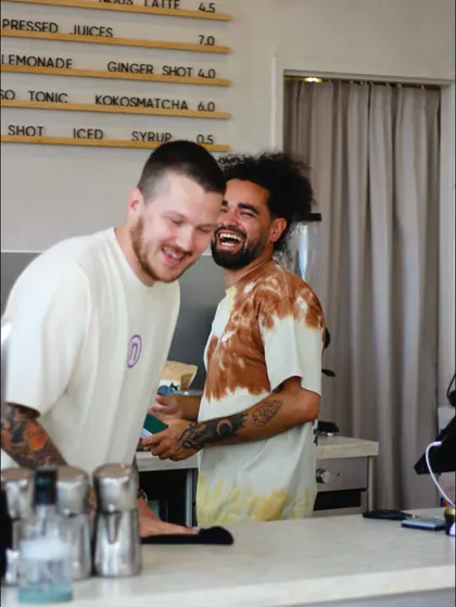 Two nous baristas laughing behind the counter under the letterboard menu.